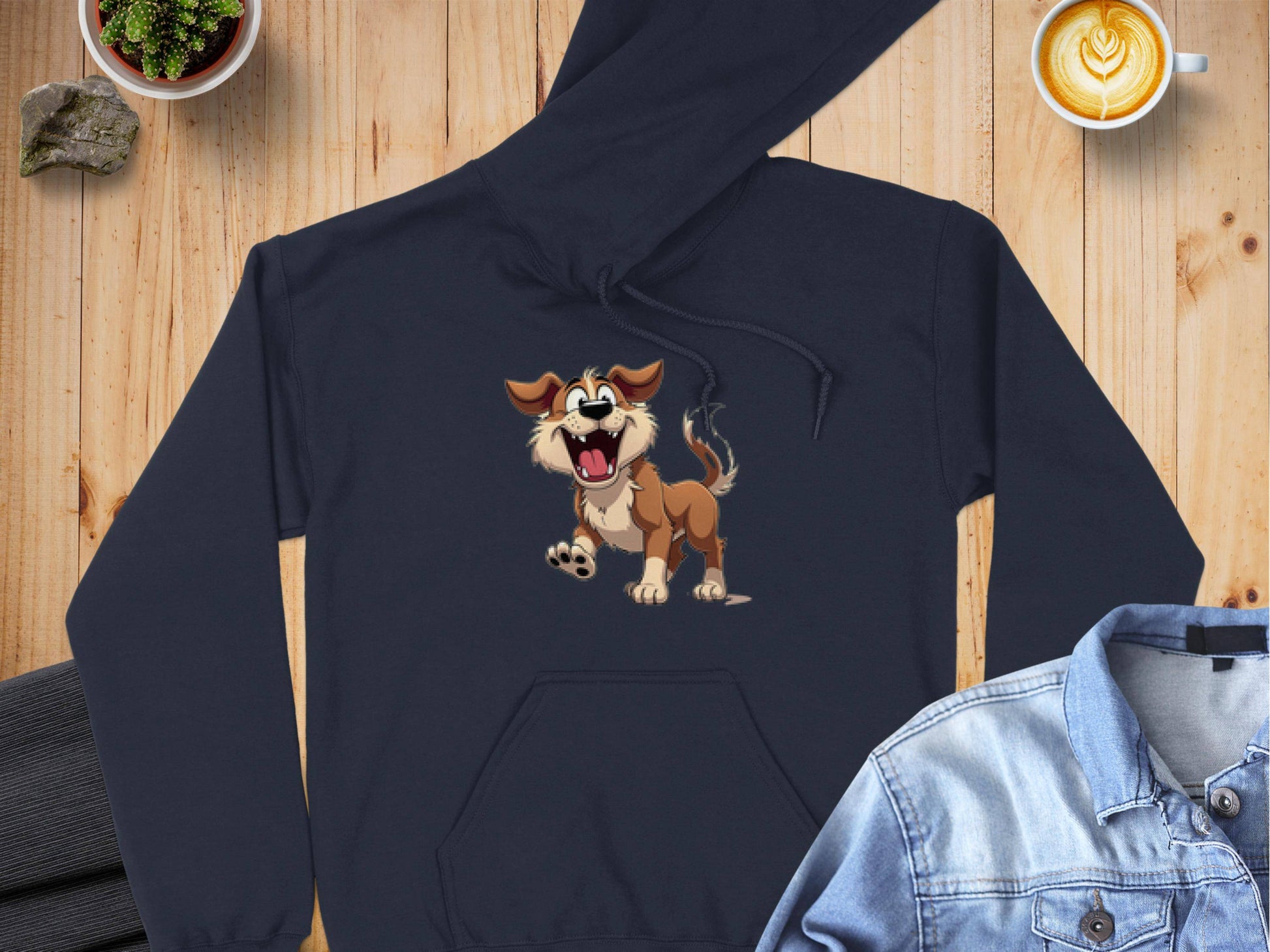 A fun wardrobe addition for dog lovers by Garment Graphics, a black cartoon puppy hoodie with a smiling, tongue-out dog is displayed on wood. Nearby are a denim jacket, latte art coffee cup, and small potted plant.