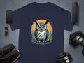 The image showcases a Garment Graphics navy blue T-shirt with an owl graphic enhanced by an orange circle. It is surrounded by a smartphone, smartwatch, wireless speaker, sneakers, and dumbbells on a gray background.