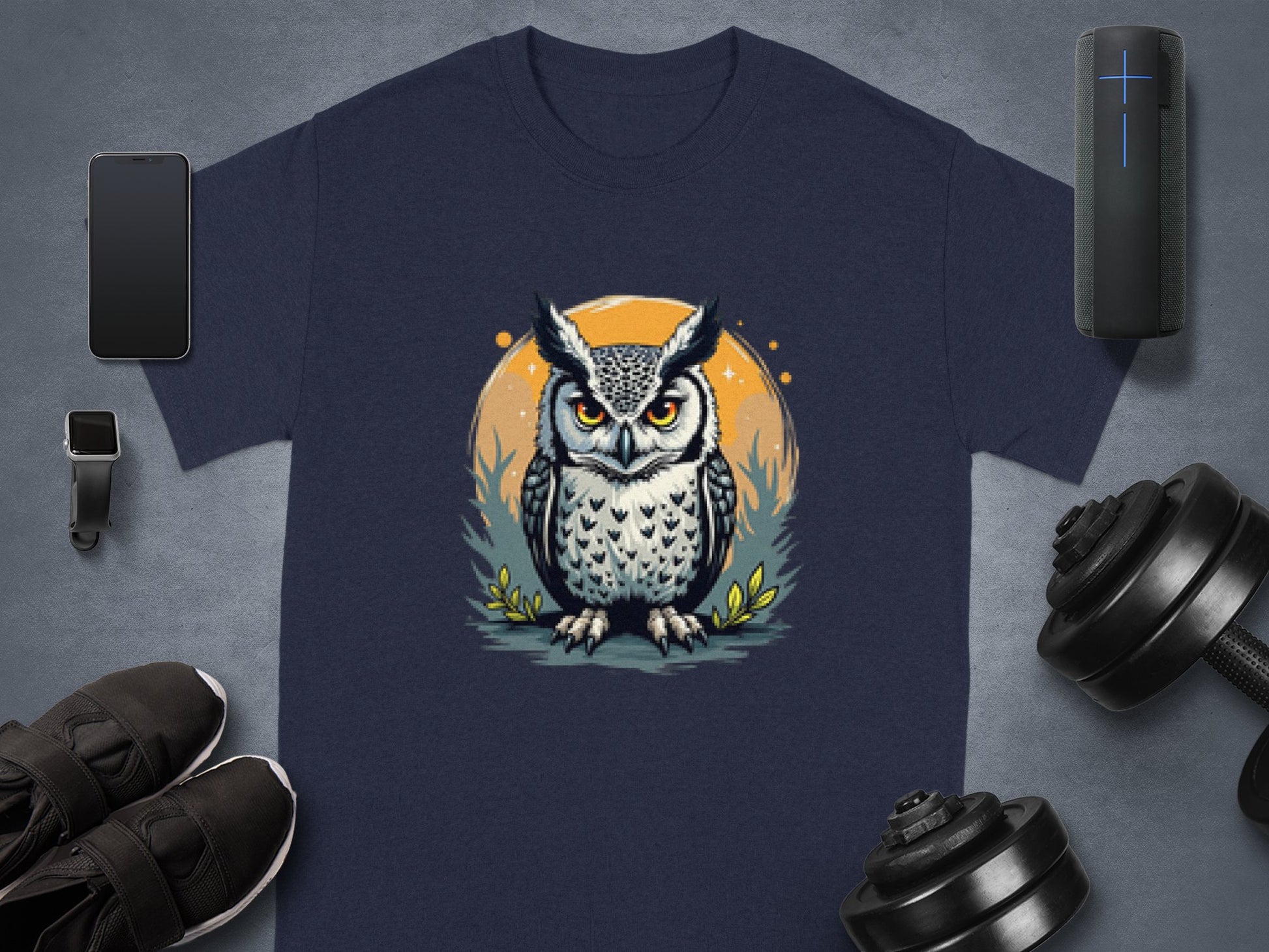 The image showcases a Garment Graphics navy blue T-shirt with an owl graphic enhanced by an orange circle. It is surrounded by a smartphone, smartwatch, wireless speaker, sneakers, and dumbbells on a gray background.