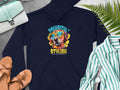 Garment Graphics presents a navy blue hoodie featuring a dog in sunglasses and headphones with Pawsitively Stylish text. Paired with a striped shirt, watch, sandals, handbag, and green fern leaves on gray—a perfect style for vibrant hoodie enthusiasts.