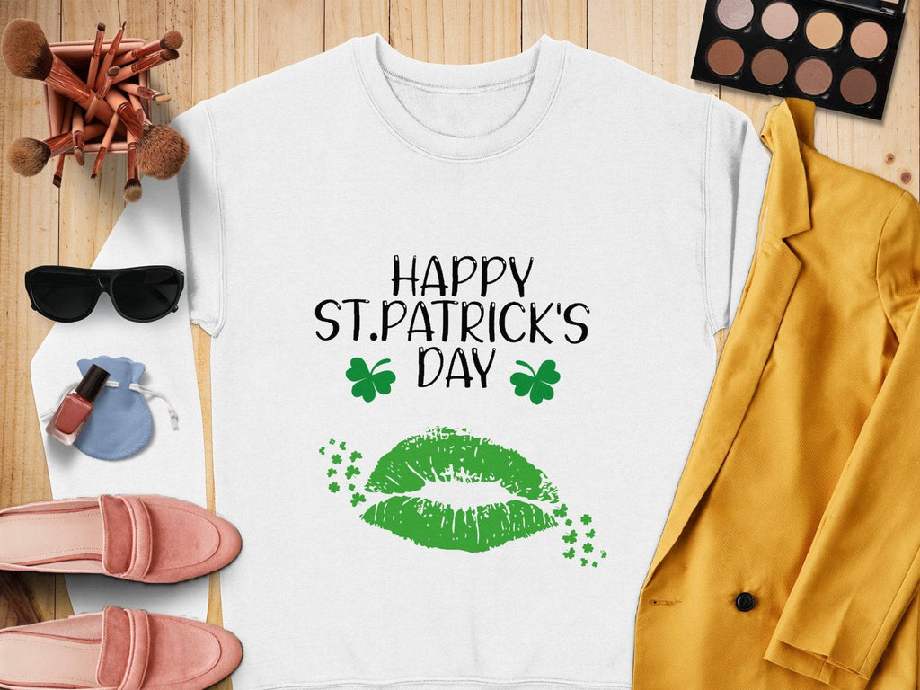 Garment Graphics white sweatshirt features HAPPY ST. PATRICKS DAY text, green lips, and shamrocks, capturing the festive spirit. Surrounded by makeup brushes, eyeshadow palette, sunglasses, lipstick, blue pouch, pink loafers, and a yellow blazer on a wooden surface.