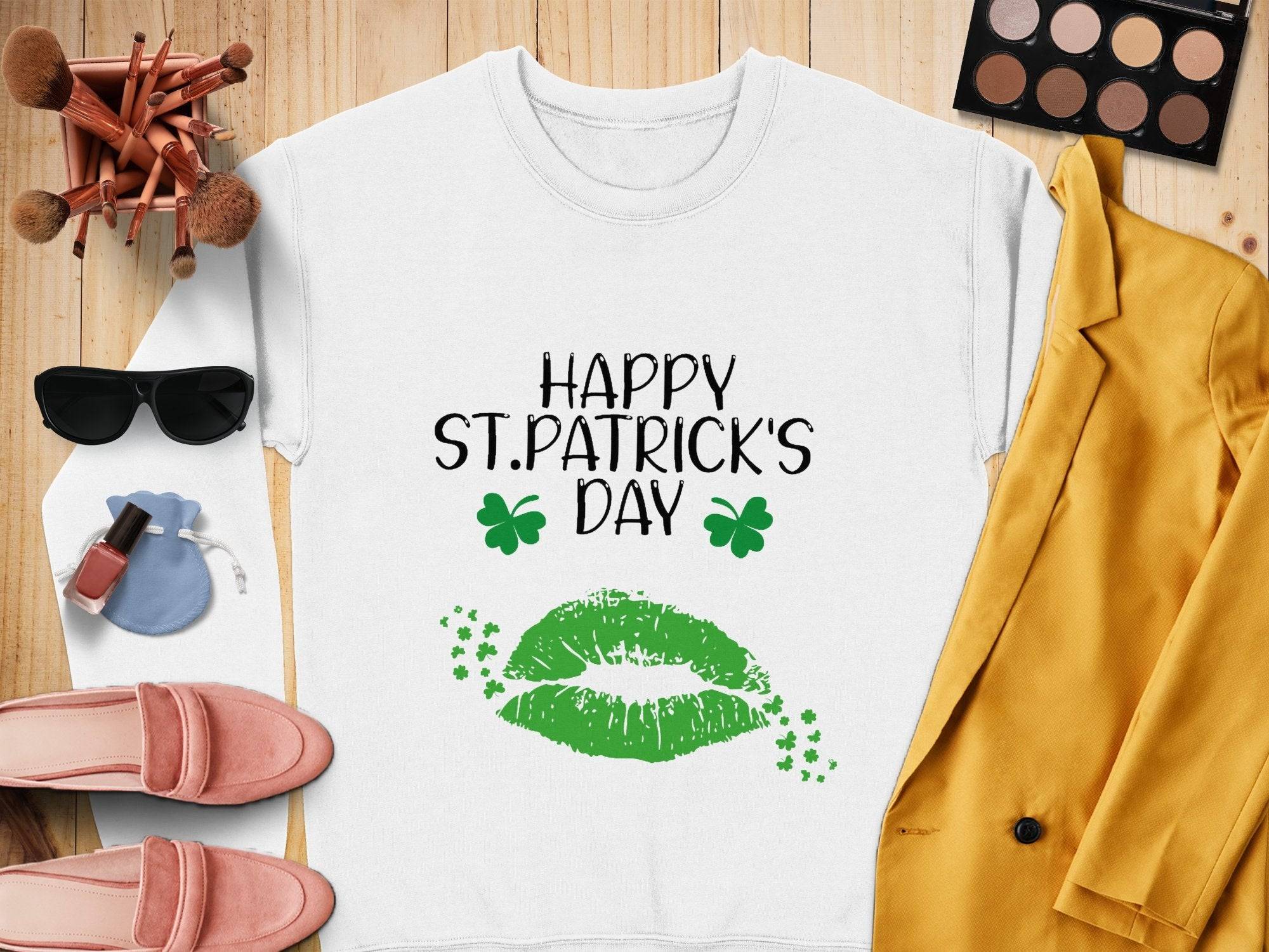 Garment Graphics white sweatshirt features HAPPY ST. PATRICKS DAY text, green lips, and shamrocks, capturing the festive spirit. Surrounded by makeup brushes, eyeshadow palette, sunglasses, lipstick, blue pouch, pink loafers, and a yellow blazer on a wooden surface.