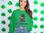 A woman beams at the camera in a green graphic crewneck sweatshirt featuring a Scania logo by Garment Graphics. She stands against a white wall with green four-leaf clovers, wearing denim shorts, reflecting her passion as a truck enthusiast.