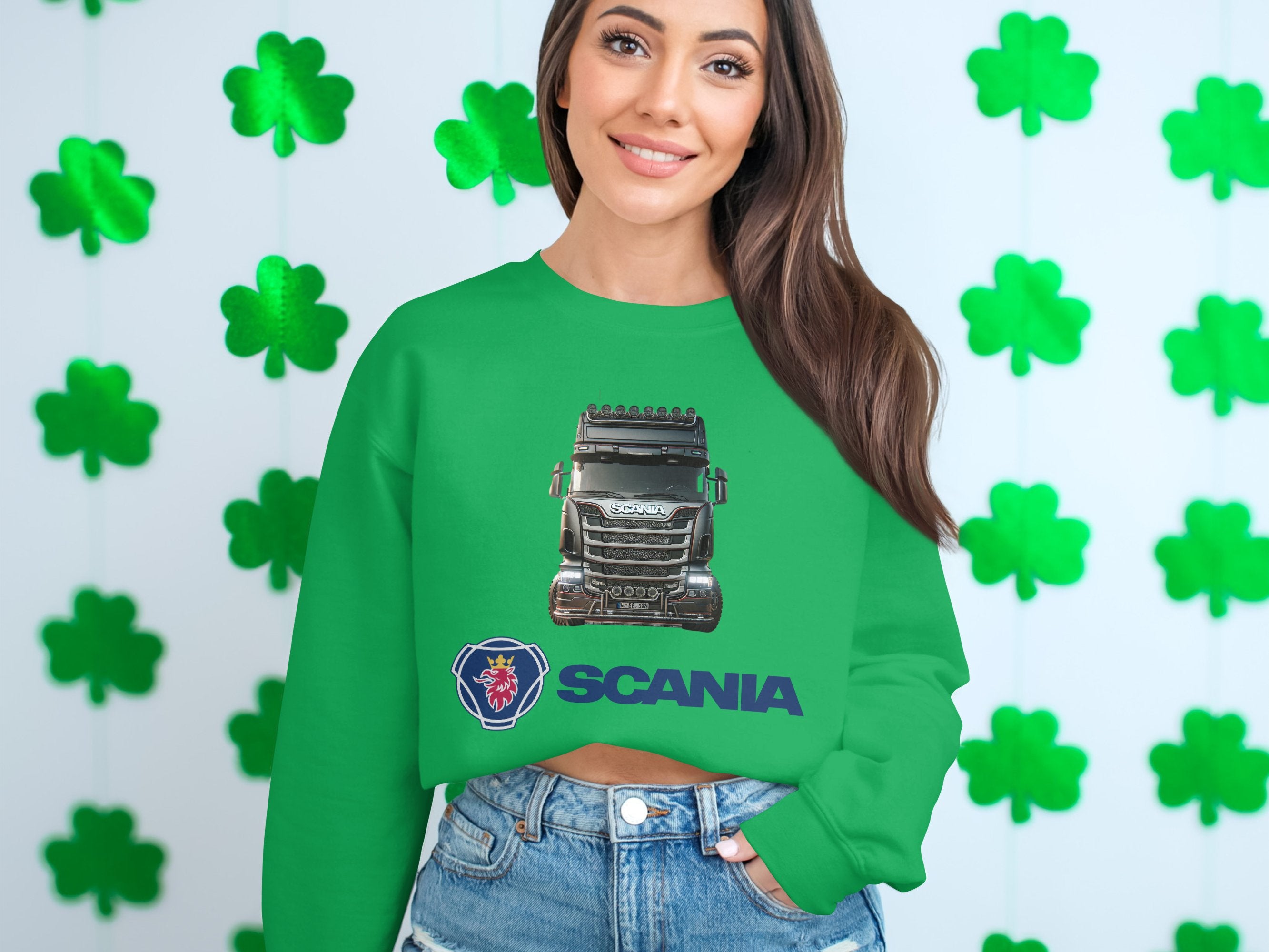 A woman beams at the camera in a green graphic crewneck sweatshirt featuring a Scania logo by Garment Graphics. She stands against a white wall with green four-leaf clovers, wearing denim shorts, reflecting her passion as a truck enthusiast.