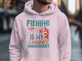 Someone wears a light pink hoodie by Garment Graphics from the Anglers Wardrobe collection, with Fishing is my anger management in blue and orange. A fish graphic completes the look, set against a blurred urban street scene.