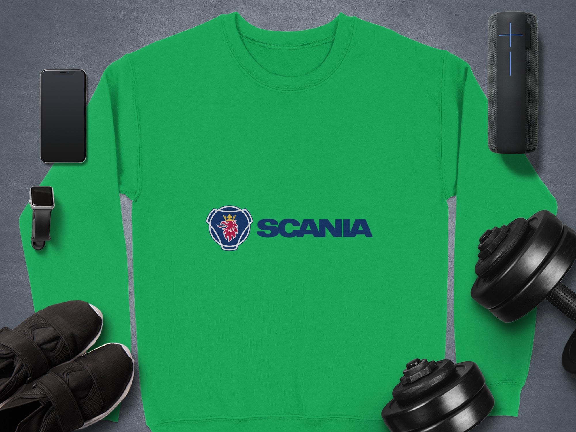 A green Garment Graphics Scania logo sweatshirt for casual outings displays the blue stylized coat of arms and SCANIA, surrounded by a smartphone, smartwatch, speaker, sleek black sneakers, and two dumbbells on a gray surface. Its designed in unisex sizing for ultimate comfort.