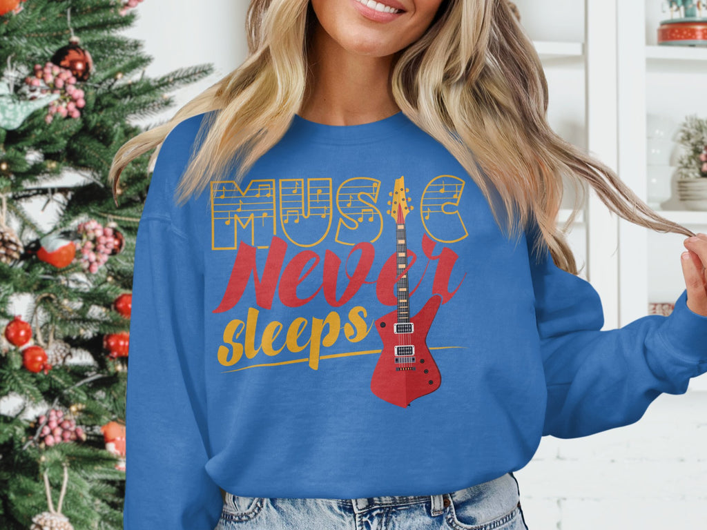 Wearing a stylish blue Music Never Sleeps Guitar sweatshirt by Garment Graphics, featuring a red electric guitar graphic, someone stands proudly by a decorated Christmas tree and bookshelf—ideal for music lovers embracing festive flair.