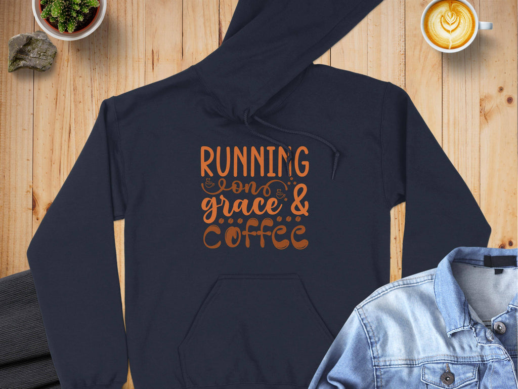 A Garment Graphics dark, medium-heavy fabric hoodie with Running on grace & coffee in orange rests on a wooden surface. Nearby are latte art coffee, a denim jacket, and a potted plant. This classic fit hoodie is ideal for cozy mornings.