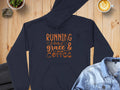 A Garment Graphics dark, medium-heavy fabric hoodie with Running on grace & coffee in orange rests on a wooden surface. Nearby are latte art coffee, a denim jacket, and a potted plant. This classic fit hoodie is ideal for cozy mornings.