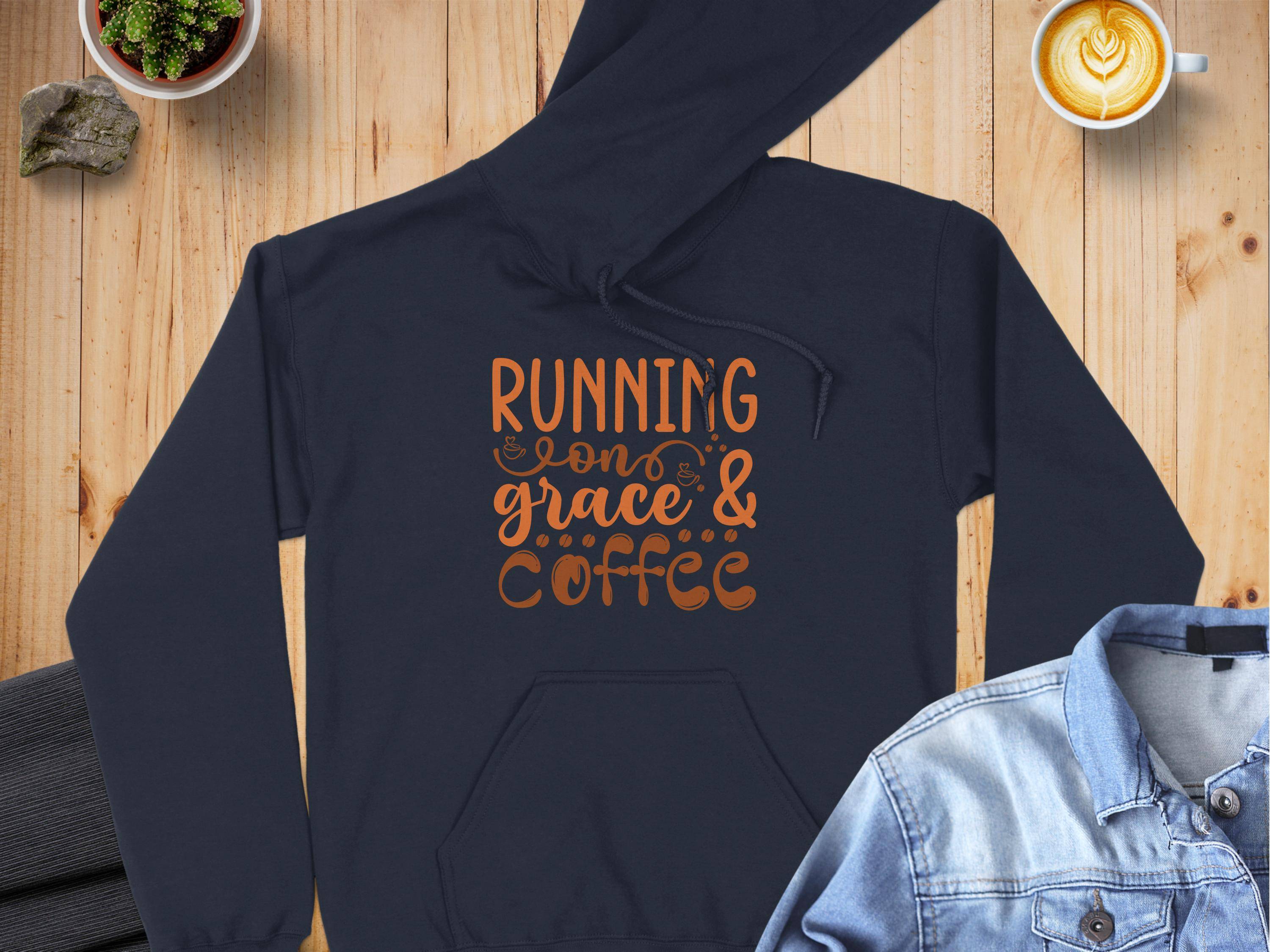 A Garment Graphics dark, medium-heavy fabric hoodie with Running on grace & coffee in orange rests on a wooden surface. Nearby are latte art coffee, a denim jacket, and a potted plant. This classic fit hoodie is ideal for cozy mornings.