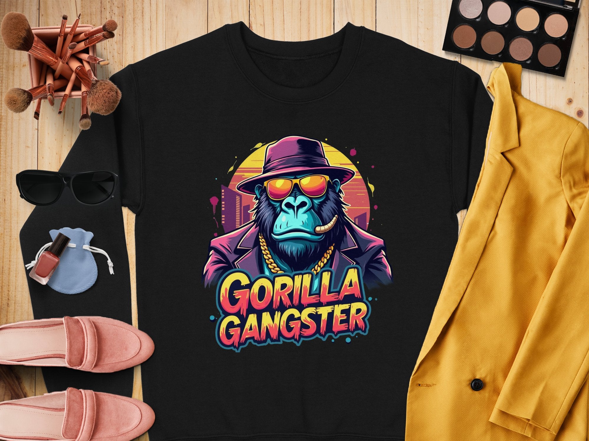 This urban black sweatshirt by Garment Graphics features a Gorilla Gangster in a hat and sunglasses, smoking a cigar, with makeup items, chic sunglasses, pink shoes, and a yellow blazer on a wooden surface. Perfect for adding edgy style to your wardrobe.