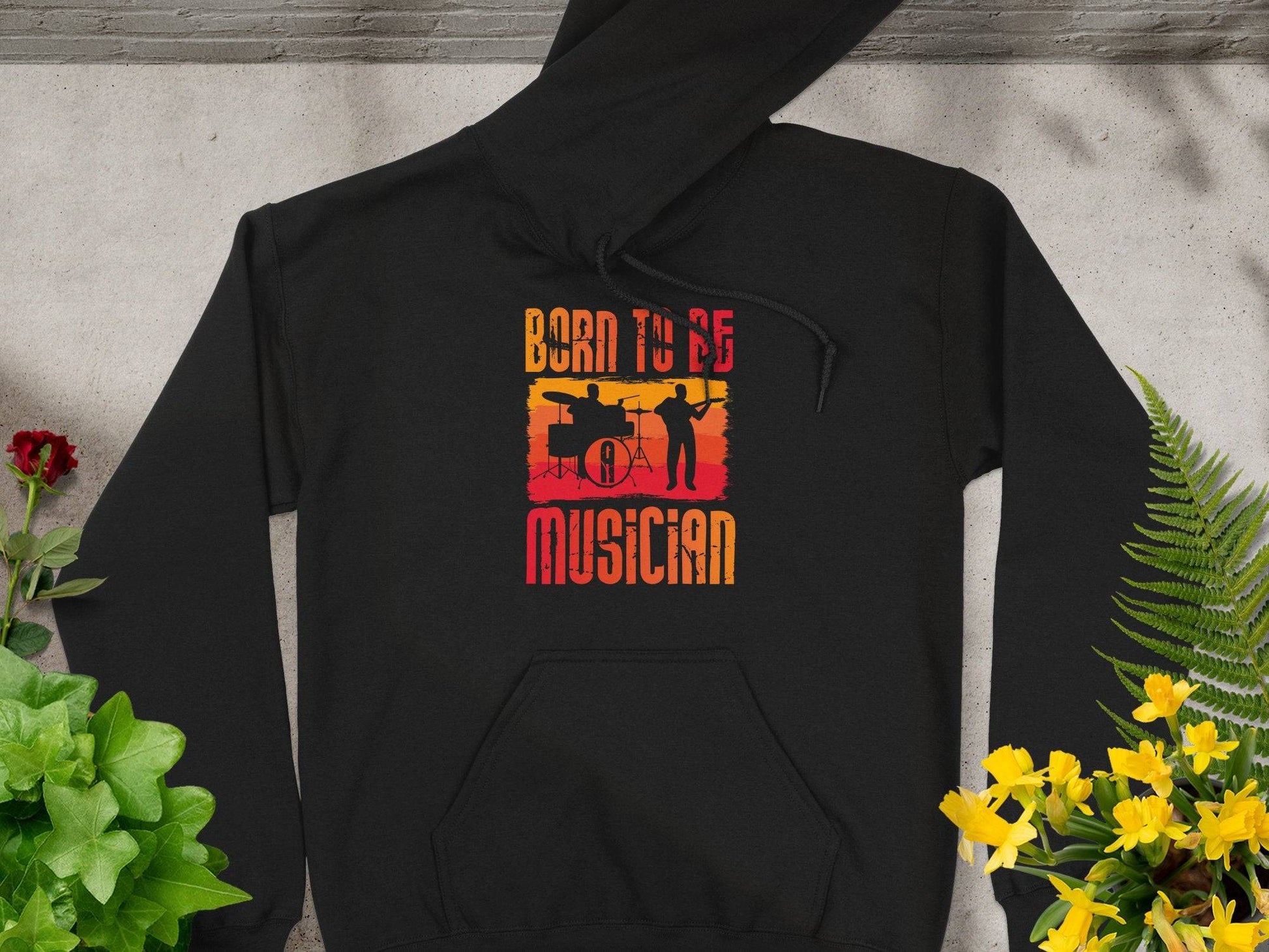 A black unisex fashion hoodie by Garment Graphics lies on concrete, adorned with a sunset gradient and musician silhouettes. Perfect for music lovers, it features the text Born to be a Musician, framed by green and yellow plants.