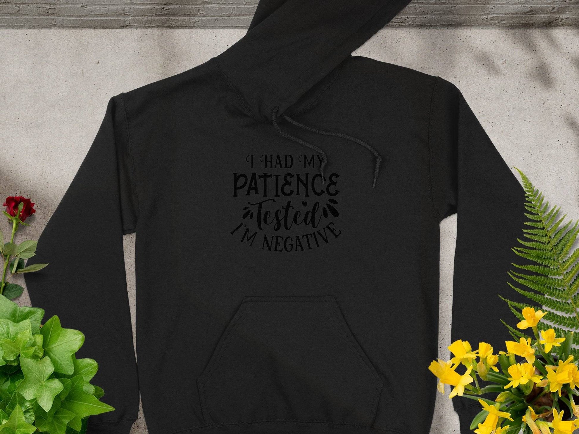 The Garment Graphics Patience Tested Hoodie features I Had My Patience Tested Im Negative, with a classic fit and medium-heavy fabric. Placed on concrete, vibrant greenery with flowers thrives to the right, while a red and green potted plant sits to the left.