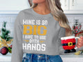 In a festive kitchen, a person wears Garment Graphics sweatshirt stating, MINE IS SO BIG I HAVE TO USE BOTH HANDS, and holds a Santas pants and boots mug, enhancing the holiday spirit.