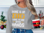 In a festive kitchen, a person wears Garment Graphics sweatshirt stating, MINE IS SO BIG I HAVE TO USE BOTH HANDS, and holds a Santas pants and boots mug, enhancing the holiday spirit.