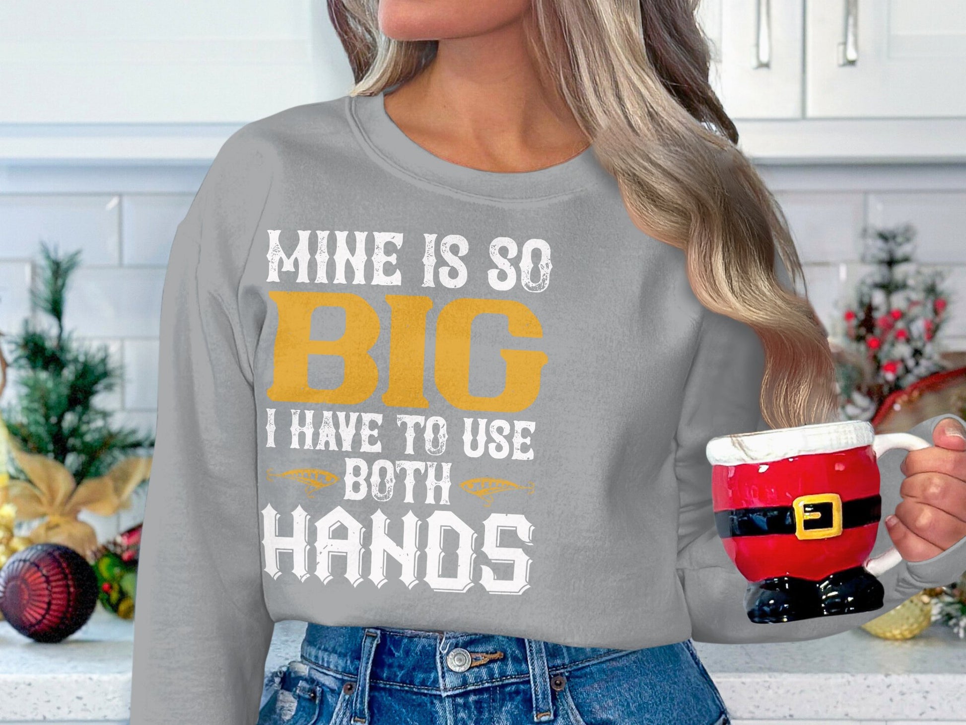 In a festive kitchen, a person wears Garment Graphics sweatshirt stating, MINE IS SO BIG I HAVE TO USE BOTH HANDS, and holds a Santas pants and boots mug, enhancing the holiday spirit.