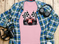 A Garment Graphics pink t-shirt with a red racing car and checkered flags is styled over a blue and green plaid shirt. Nearby, a black camera, headphones, and a takeaway coffee cup on the wood surface complete the motorsport enthusiasts setup.