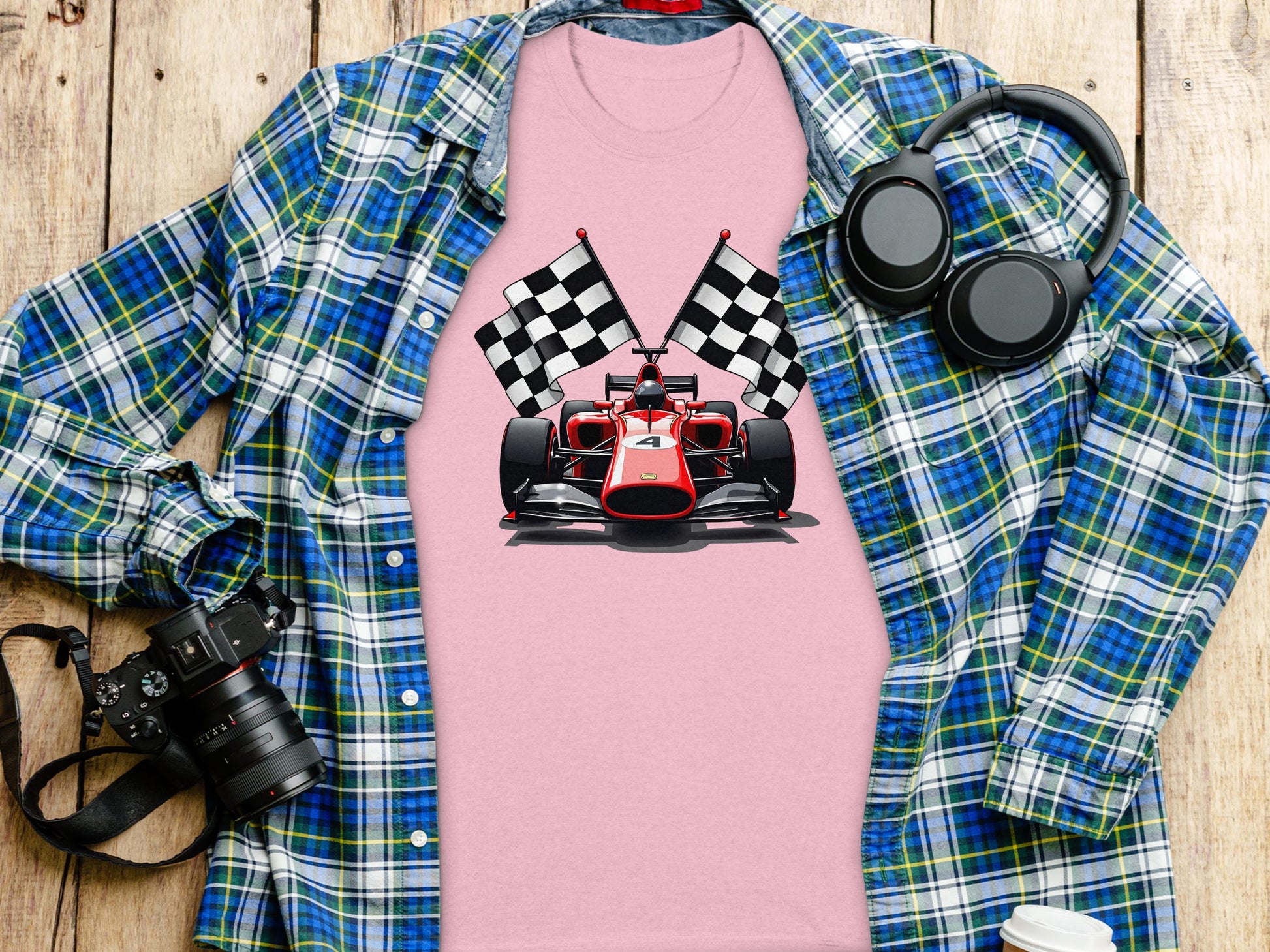 A Garment Graphics pink t-shirt with a red racing car and checkered flags is styled over a blue and green plaid shirt. Nearby, a black camera, headphones, and a takeaway coffee cup on the wood surface complete the motorsport enthusiasts setup.