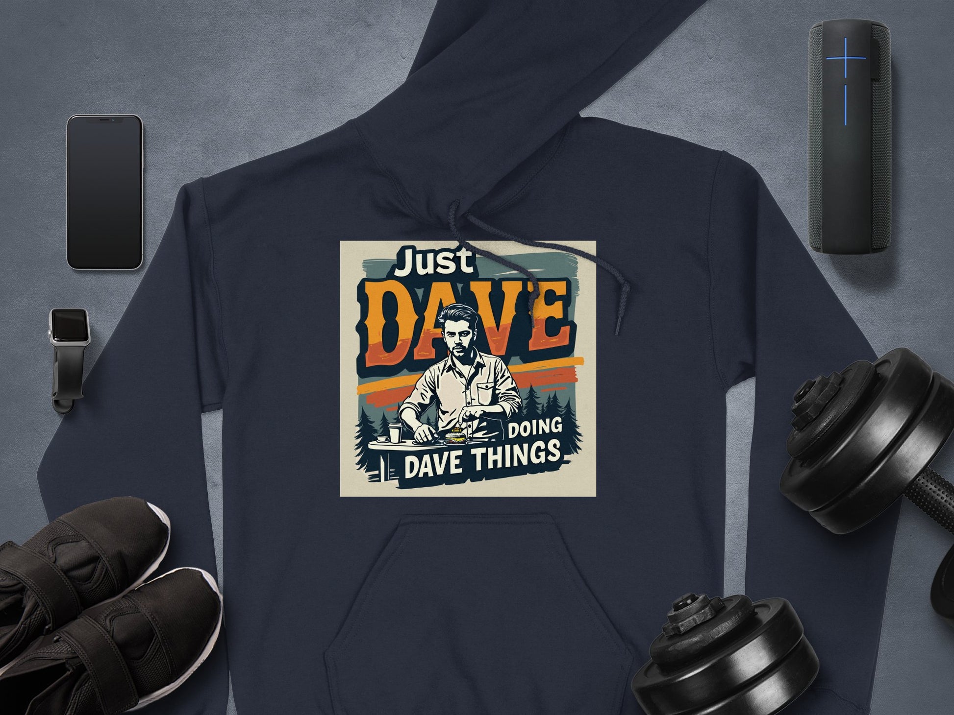 The Garment Graphics hoodie features Just Dave Doing Dave Things with an illustration of a man. Made from medium-heavy fabric, it lies on a gray surface with a smartphone, smartwatch, wireless speaker, sneakers, and dumbbells.