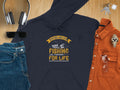 A Garment Graphics navy fishing hoodie with the Father and Son Fishing Partners for Life design rests on a wooden surface. Nearby, jeans, sunglasses, headphones, a watch, and keys enhance the display. An orange shirt with glasses tops it off—ideal for fishing enthusiasts.