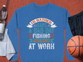 On a brown surface lies a Garment Graphics fishing sweatshirt with the slogan Id rather have a bad day fishing than a good day at work, ideal for enthusiasts. Nearby are unisex essentials: a water bottle, gray towel, black shorts, and an orange basketball.