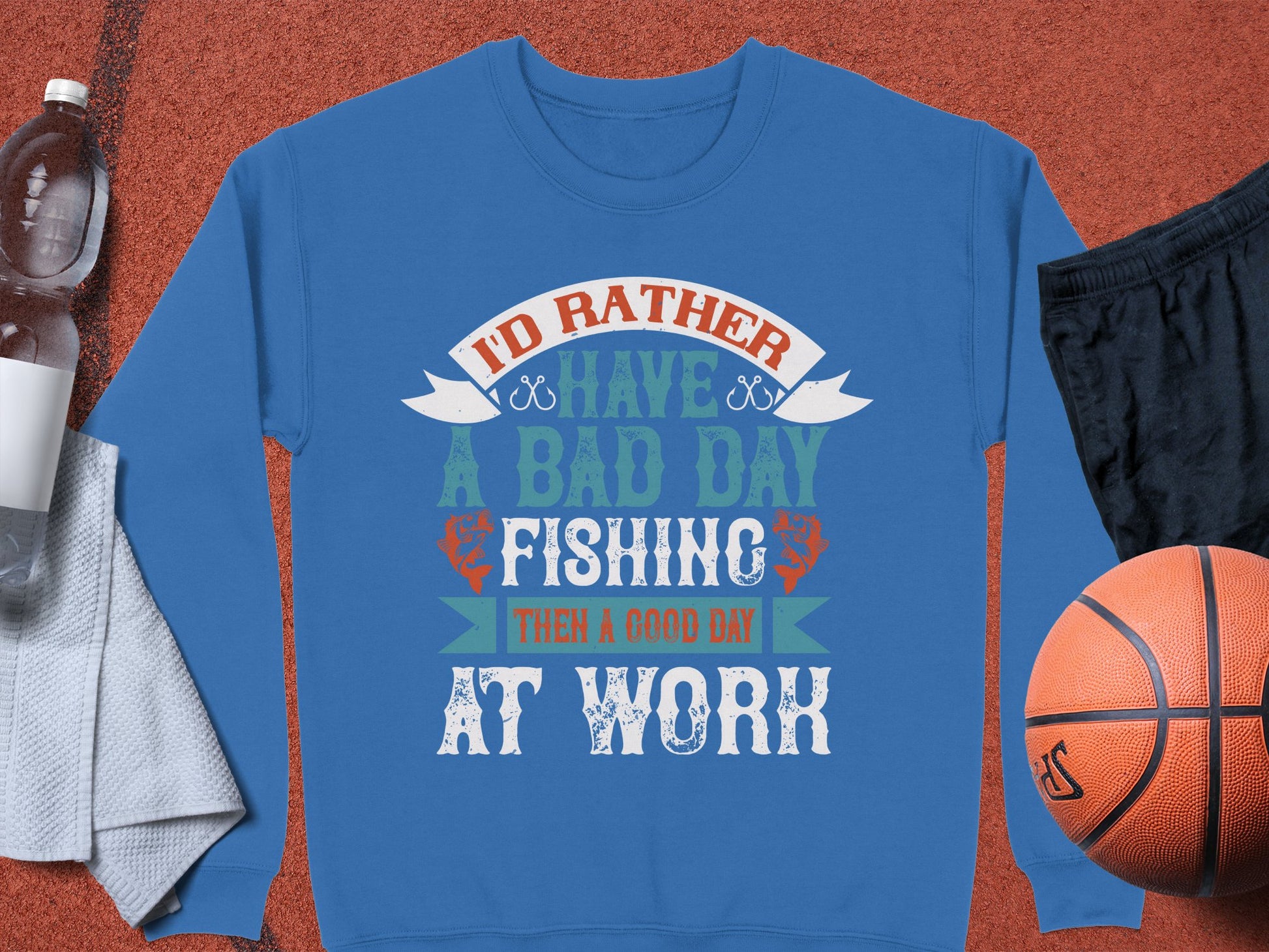 On a brown surface lies a Garment Graphics fishing sweatshirt with the slogan Id rather have a bad day fishing than a good day at work, ideal for enthusiasts. Nearby are unisex essentials: a water bottle, gray towel, black shorts, and an orange basketball.