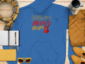 A classic fit blue hoodie by Garment Graphics, featuring Music Never Sleeps in yellow and red with a guitar design, lies on a wooden surface. It pairs well with a yellow scarf, quilted jacket, shoes, sunglasses, notebook, and coffee cup.