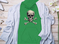 A vibrant green Garment Graphics hoodie with a skull and crossbones design rests on a light wooden surface. Its accented by a light gray cardigan, chic white heels, elegant jewelry, and a vase of stunning white flowers.