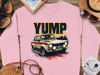 This pink sweatshirt by Garment Graphics features a rally car design with YUMP above it, showcasing a vintage car with four headlights and a red stripe, surrounded by a hat, shoes, notebook, sunglasses, and camera on a wooden surface.