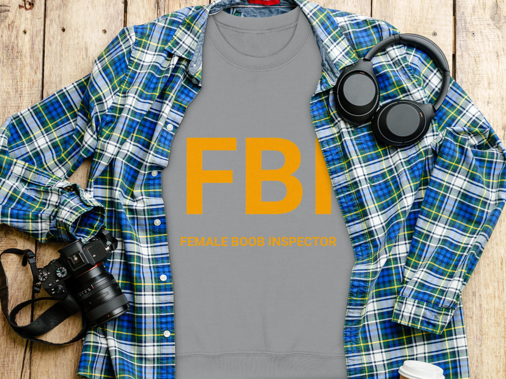 A grey sweatshirt from Garment Graphics with FBI in bold orange letters above “Female Boob Inspector” is shown on a wooden surface, surrounded by a blue plaid shirt, headphones, a camera, and a coffee cup—great for cheeky humor fans in unisex sizing.