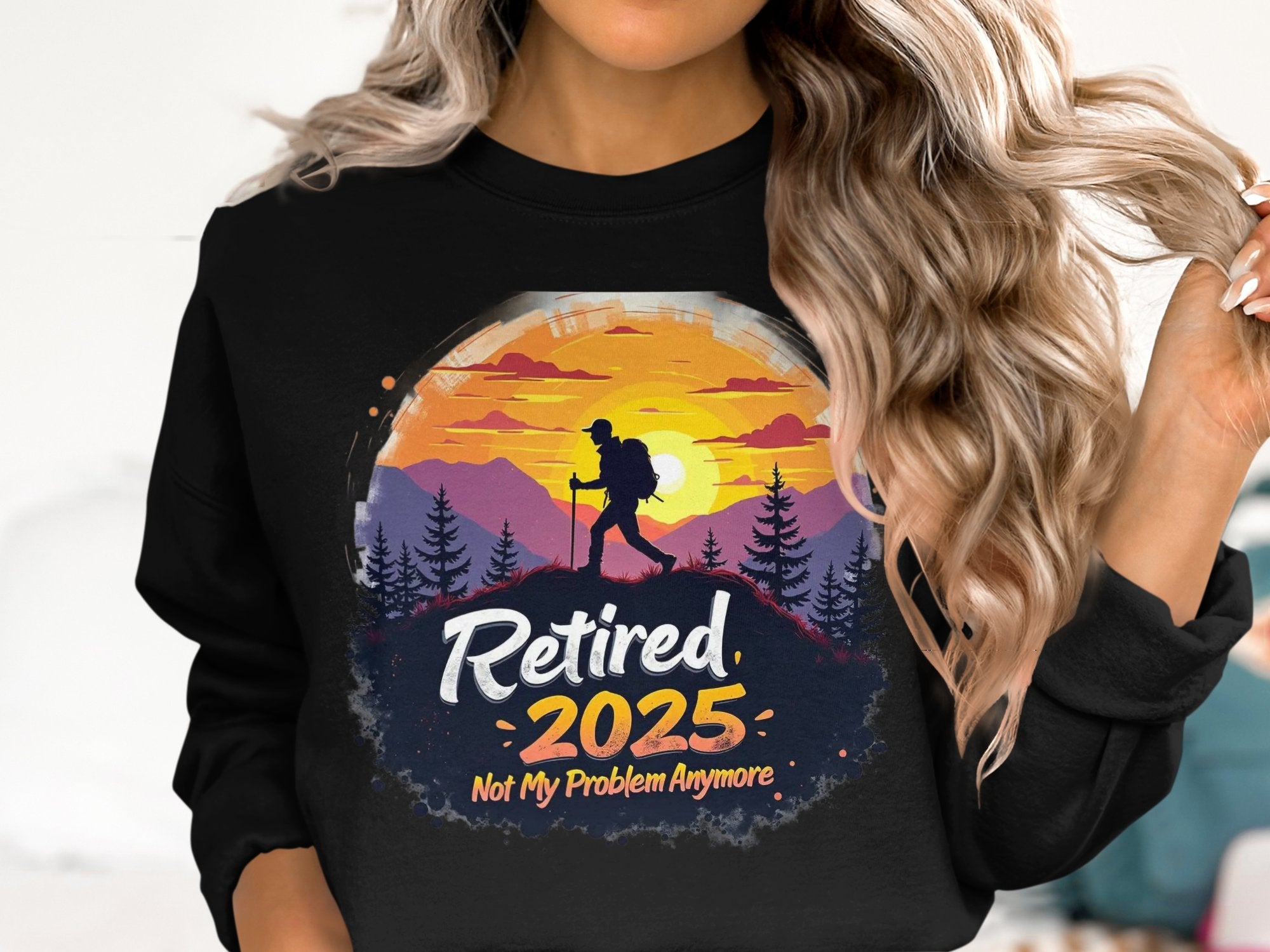 A person with long, wavy blonde hair dons a black “Retired 2025 Not My Problem Anymore” unisex sweatshirt by Garment Graphics, featuring a colorful hiking design against a sunset.