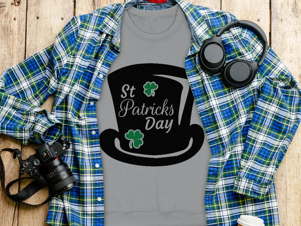 A Garment Graphics grey t-shirt featuring a black top hat and green shamrocks with St Patricks Day text captures the festive spirit beneath a blue and green plaid shirt. Nearby, a camera, headphones, and coffee cup sit on a wooden surface.