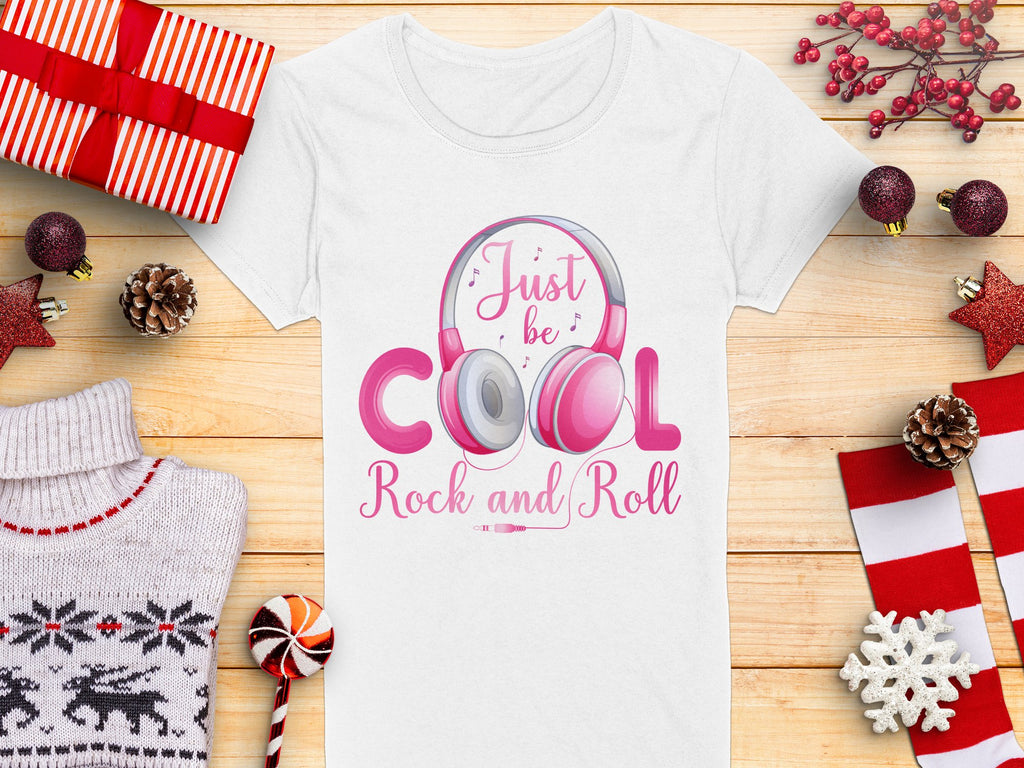 The Garment Graphics white headphones t-shirt displays a pink headphones design with Just be cool, Rock and Roll text. Its ideal for music lovers, shown among wrapped gifts, pine cones, red stars, a candy cane, a red-and-white sweater, and red-berried sprigs on wood.