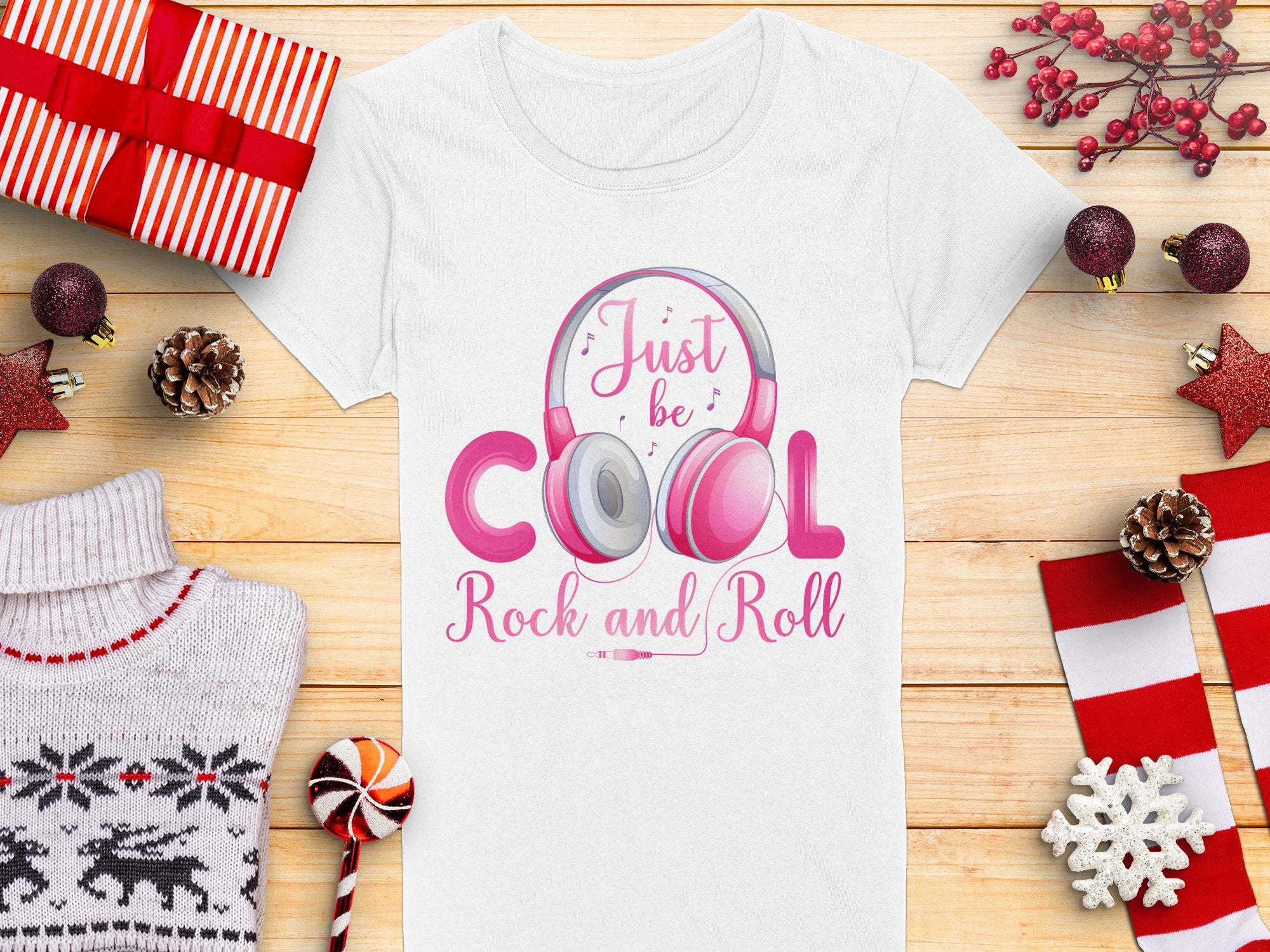 The Garment Graphics white headphones t-shirt displays a pink headphones design with Just be cool, Rock and Roll text. Its ideal for music lovers, shown among wrapped gifts, pine cones, red stars, a candy cane, a red-and-white sweater, and red-berried sprigs on wood.