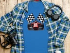 A Garment Graphics blue T-shirt with a racing car and checkered flags graphic is displayed on a wooden surface, paired with a plaid shirt, camera, headphones, and white cup—an ideal tribute to the excitement of motorsport.