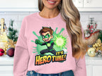 A woman stands in the kitchen wearing a Garment Graphics cartoon-themed pink sweatshirt, showcasing a green-glowing superhero and the phrase Its Hero Time! Her wavy long hair complements the festive holiday ornaments on the kitchen counter.
