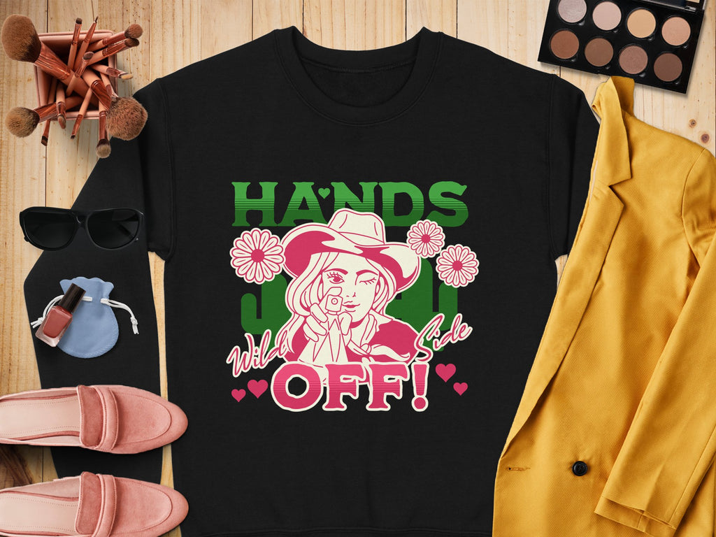 Flat lay featuring Garment Graphics’ black t-shirt with HANDS OFF! and a cartoon cowboy design, surrounded by a yellow jacket, sunglasses, pink shoes, makeup palette, brushes, and nail polish on a wooden surface. Ideal for those who love unique designs and loose fits.