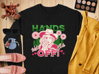 Flat lay featuring Garment Graphics’ black t-shirt with HANDS OFF! and a cartoon cowboy design, surrounded by a yellow jacket, sunglasses, pink shoes, makeup palette, brushes, and nail polish on a wooden surface. Ideal for those who love unique designs and loose fits.