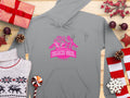 A light gray hoodie featuring a pink Beach Girl graphic with three silhouettes of women in swimsuits by Garment Graphics, surrounded by wrapped gifts, pinecones, Christmas decorations, a white reindeer sweater, and a round lollipop on wood. Embrace casual style with Always Party Beach Girl Hoodies.