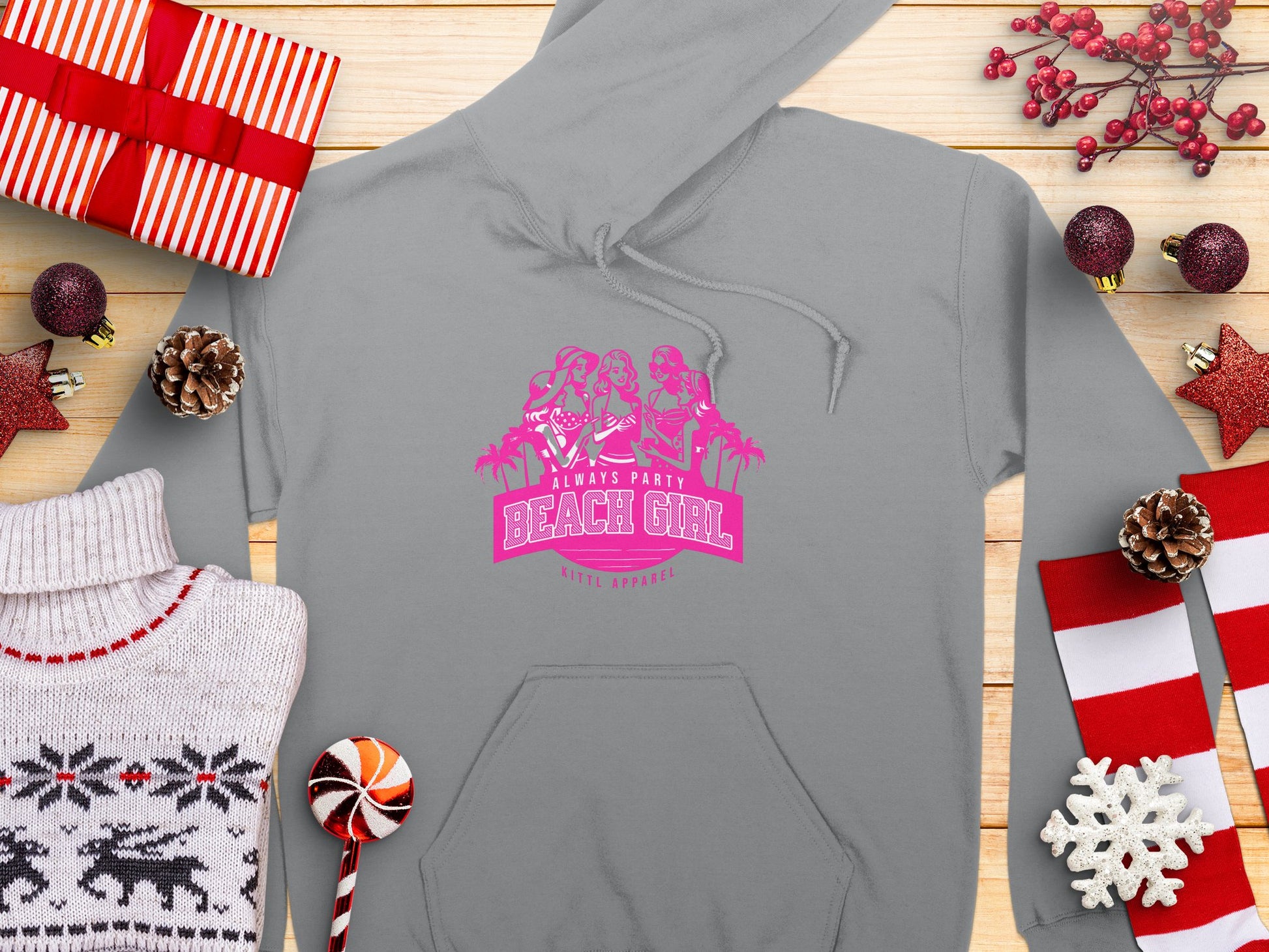 A light gray hoodie featuring a pink Beach Girl graphic with three silhouettes of women in swimsuits by Garment Graphics, surrounded by wrapped gifts, pinecones, Christmas decorations, a white reindeer sweater, and a round lollipop on wood. Embrace casual style with Always Party Beach Girl Hoodies.