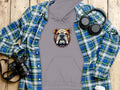 A gray hoodie from Garment Graphics, featuring a bold bulldog logo, stands out against a blue and green plaid shirt. Headphones, a camera, and a white takeaway cup complete the ensemble on the wooden surface.