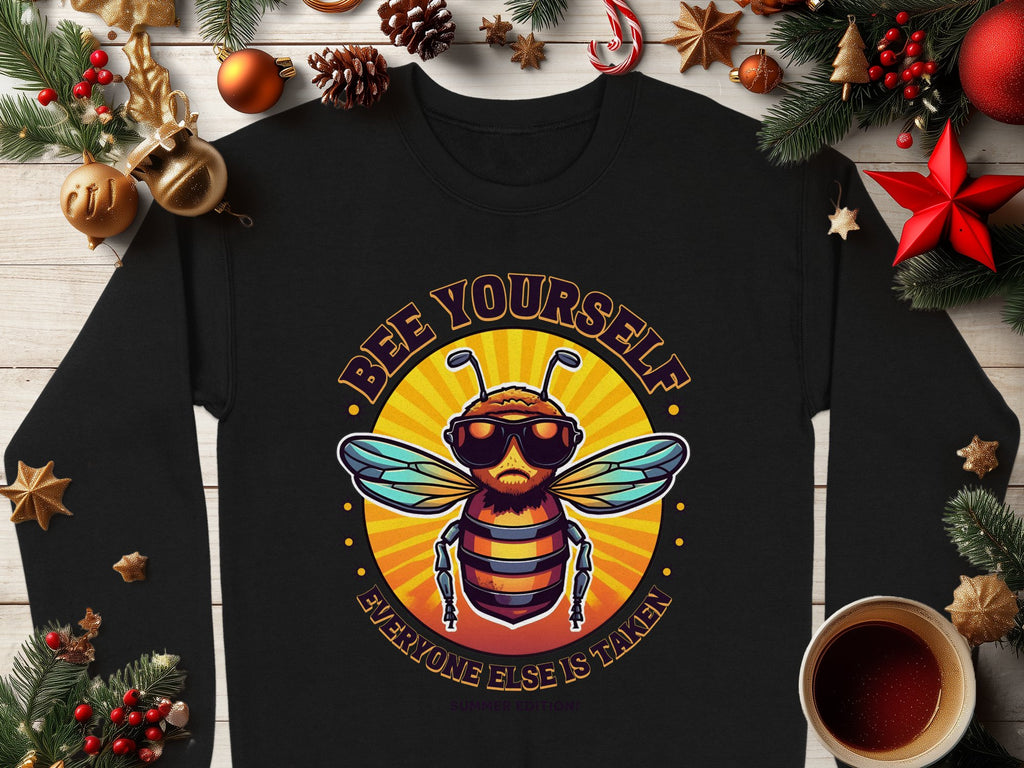 Flat lay of a Garment Graphics black shirt displaying a bee with sunglasses and the text Bee Yourself above and Everyone Else Is Taken below in retro font. Ideal for unisex sizing, its surrounded by holiday decorations and a coffee cup on a wooden surface.