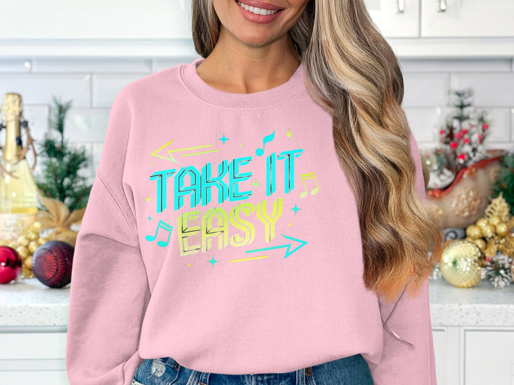 In a holiday-themed kitchen decorated with ornaments and Christmas trees, a woman sports casual wear—a Garment Graphics unisex pink sweatshirt with Take It Easy in colorful letters. Her long, wavy hair completes the picture.