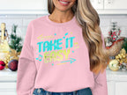 In a holiday-themed kitchen decorated with ornaments and Christmas trees, a woman sports casual wear—a Garment Graphics unisex pink sweatshirt with Take It Easy in colorful letters. Her long, wavy hair completes the picture.