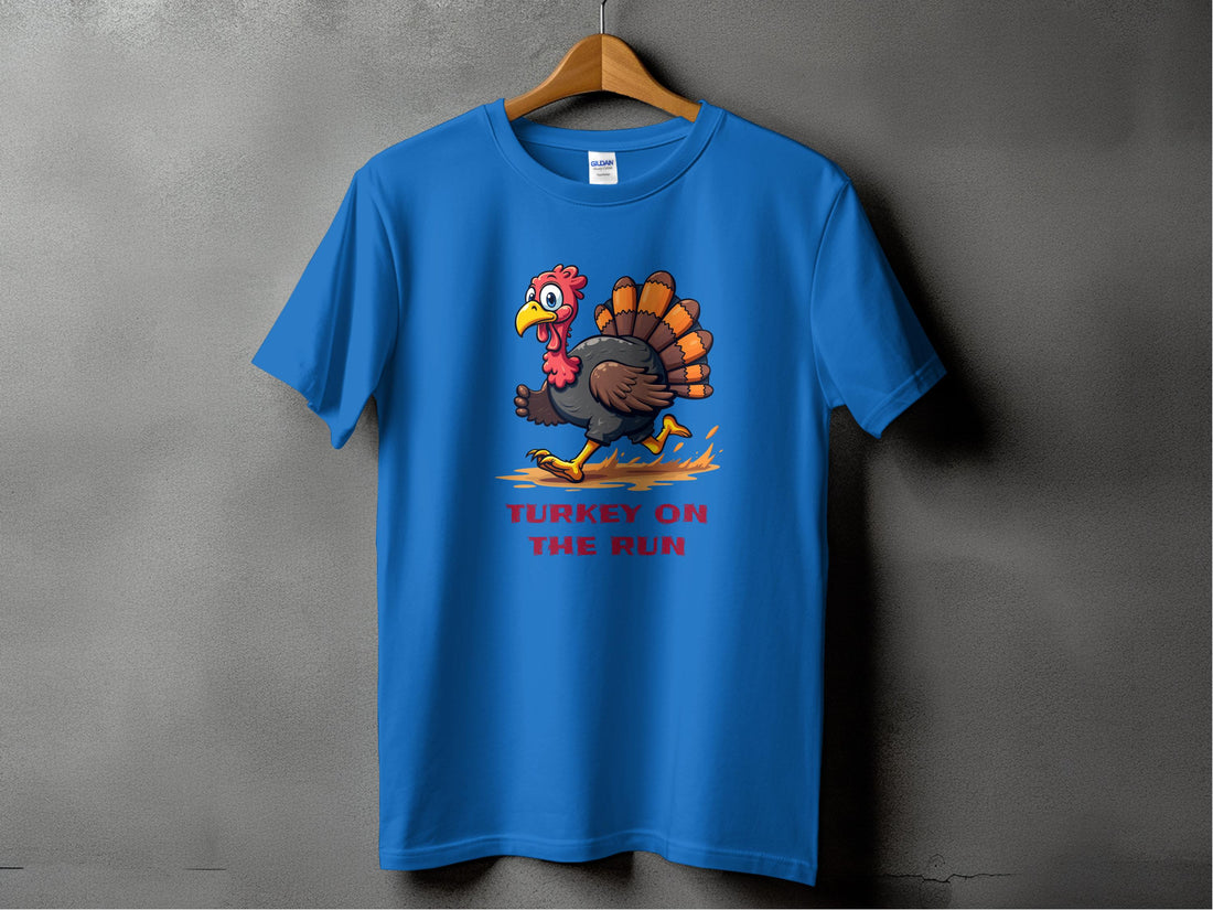 A blue T-shirt by Garment Graphics hangs on a wooden hanger, showcasing a funny cartoon turkey running over TURKEY ON THE RUN in bold red letters. Ideal for fans of witty designs, the graphic stands out against the textured gray wall.