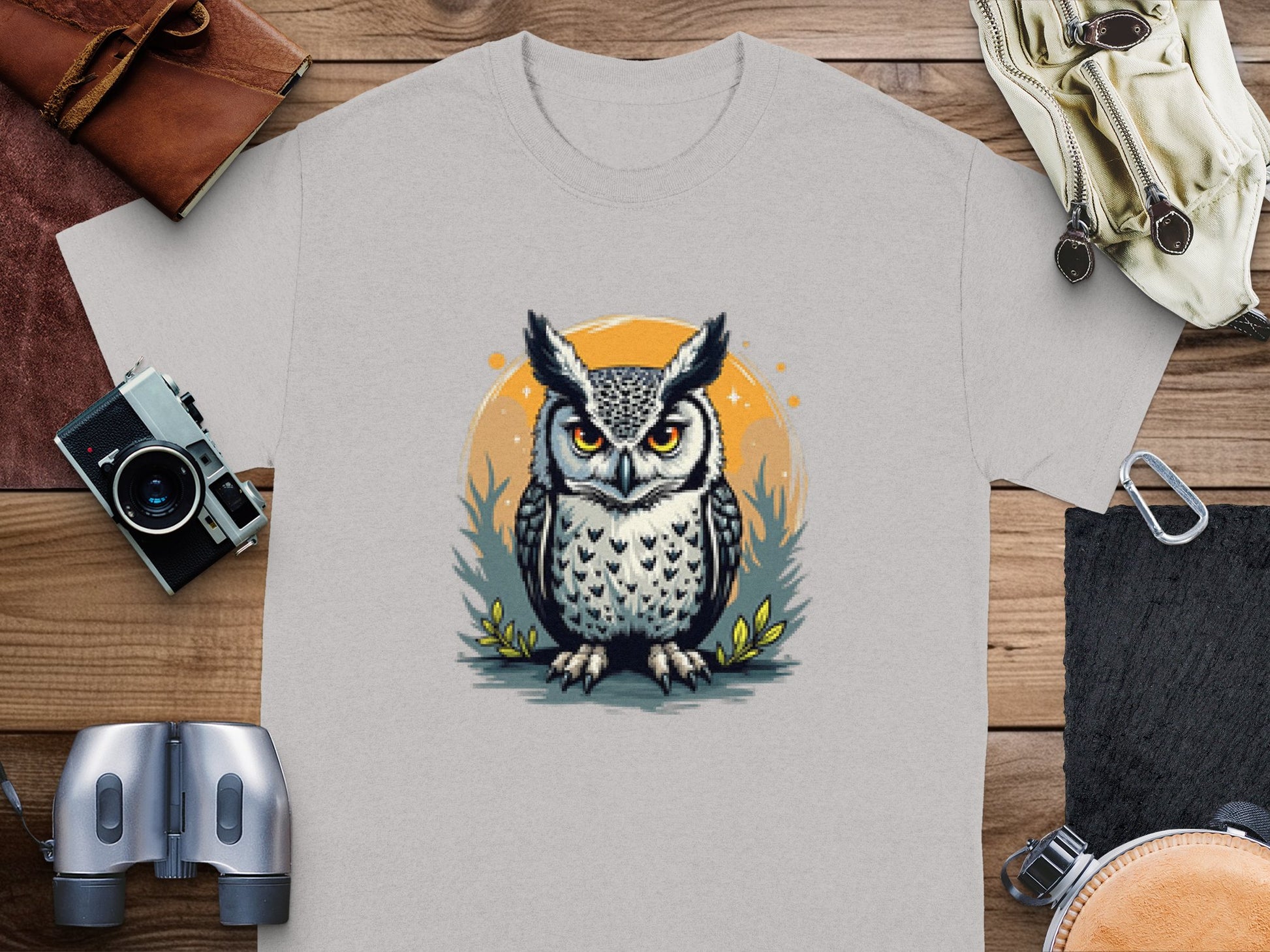 A light gray T-shirt by Garment Graphics features an owl with large eyes and a serious expression, encircled by grass and an orange ring. Laid on a wooden surface with a camera and binoculars nearby, this tee blends artistic flair and practicality.
