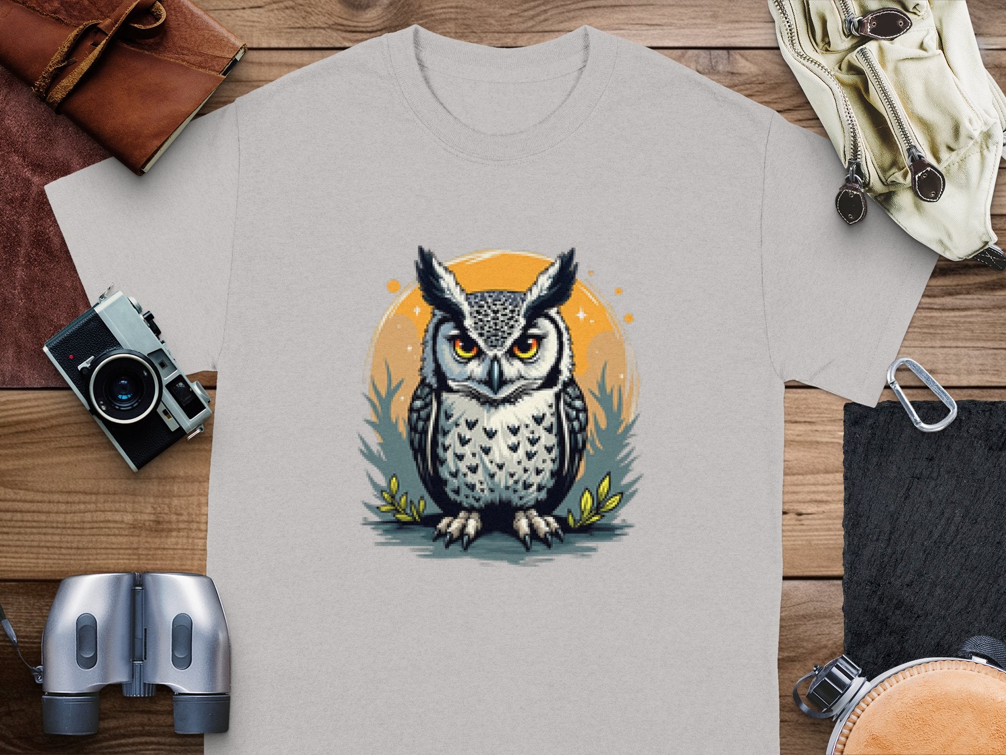 A light gray T-shirt by Garment Graphics features an owl with large eyes and a serious expression, encircled by grass and an orange ring. Laid on a wooden surface with a camera and binoculars nearby, this tee blends artistic flair and practicality.