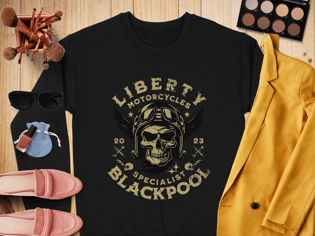 Garment Graphics offers a black T-shirt featuring a skull with goggles and Liberty Motorcycles Specialist Blackpool 2023. Pair it with a mustard blazer for an edgy biker style, and accessorize with pink loafers, sunglasses, makeup brushes, nail polish, and an eyeshadow palette.