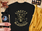 Garment Graphics offers a black T-shirt featuring a skull with goggles and Liberty Motorcycles Specialist Blackpool 2023. Pair it with a mustard blazer for an edgy biker style, and accessorize with pink loafers, sunglasses, makeup brushes, nail polish, and an eyeshadow palette.