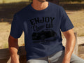 A person sits on a wooden bench wearing a navy blue t-shirt by Garment Graphics, featuring a vintage car graphic with the text Enjoy Classic Car, Vintage Ride Community, Aircooled, Est. 1980, ideal for car enthusiasts who love timeless journeys.